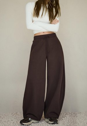 Femme portant un crop top blanc à manches longues, un pantalon large foncé avec une taille à cordon, et des baskets noires, debout sur du gravier, bras croisés.