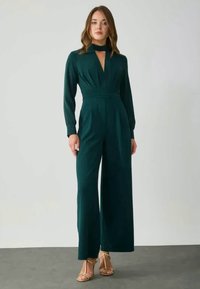 Donkergroene jumpsuit met lange mouwen, een keyhole-uitgesneden halslijn, geplooide taille en wijde pijpen. Gladde stoftextuur.