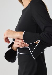 Schwarzes Kleid mit weißen Paspeln an den Ärmeln, mit ausladenden Ärmeln und einer taillierten Silhouette. Weicher Stoff mit glatter Textur.
