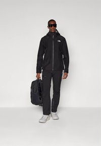 Melna ūdensizturīga jaka ar kapuci, priekšējo rāvējslēdzēju un logotipu. Atbilstošas melnas bikses un balti sporta apavi. Melna mugursoma rokā.