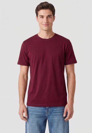 Jonge man staand en recht vooruit kijkend, draagt een effen bordeauxrood T-shirt met korte mouwen en een blauwe spijkerbroek tegen een lichtgrijze achtergrond.