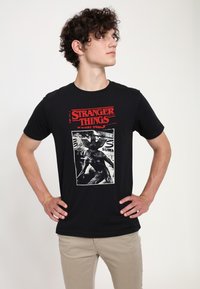 Camiseta negra de algodón con texto rojo "STRANGER THINGS" y un gráfico blanco de una criatura en el centro, de manga corta y cuello redondo.