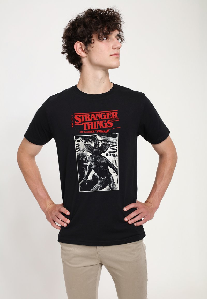 Camiseta negra de algodón con texto rojo "STRANGER THINGS" y un gráfico blanco de una criatura en el centro, de manga corta y cuello redondo.