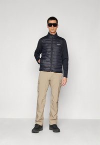 Svart vattert vest med glidelås foran, lange ermer og logo, kombinert med beige cargobukser og svarte sko. Tilbehør: mørke solbriller.