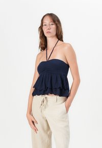 Haut sans bretelles bleu marine avec un corsage froncé, des volants en cascade à l'ourlet et de fines lacets. Porté avec un pantalon ample de couleur claire.