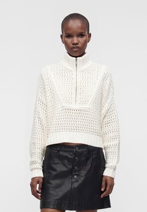 Femme aux cheveux courts portant un pull en maille blanc zippé et une jupe en cuir noire boutonnée, debout sur un fond clair uni.