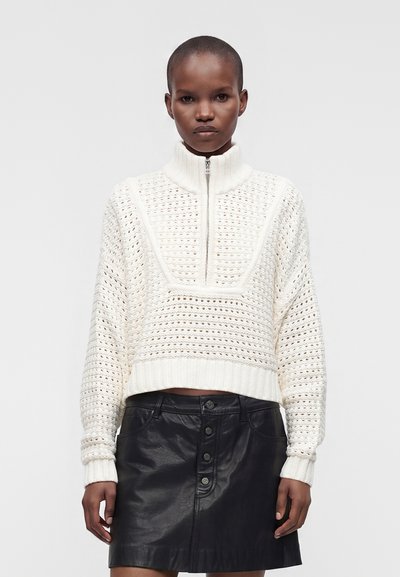 Femme aux cheveux courts portant un pull en maille blanc zippé et une jupe en cuir noire boutonnée, debout sur un fond clair uni.