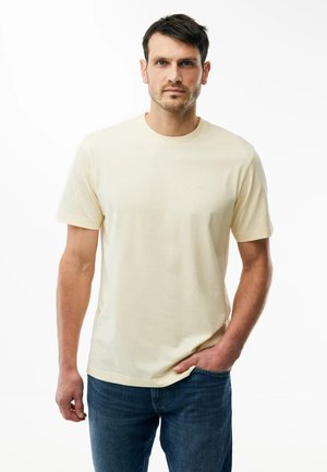 Mężczyzna o krótkich ciemnych włosach, ubrany w jasnobeżowy t-shirt z krótkim rękawem i niebieskie dżinsy, stoi z jedną ręką w kieszeni na białym tle.