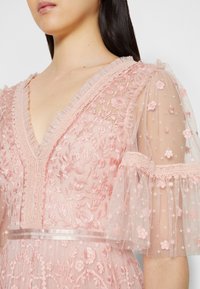 Femme portant une robe en dentelle rose avec broderies florales, décolleté en V volanté, manches bouffantes transparentes et ceinture en satin.