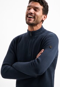 Marineblauwe geribbelde trui met een hoge kraag en een rits tot halverwege. Kenmerkt zich door een gestructureerde stof en een klein hexagonaal logo op de linker mouw.