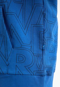 Modrá mikina s geometrickým černým liniovým vzorem, s žebrovanými manžetami a dolním lemem, a malou visačkou viditelnou na boku.