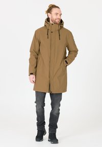 Khaki parka-jack met capuchon, ritssluiting, verstelbare trekkoorden en zijzakken, gedragen over donkere jeans en zwarte laarzen.