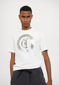 Hvid bomulds t-shirt med et cirkulært sølvlogo med slange mønster og teksten "JUST CAVALLI". Korte ærmer og afslappet pasform.