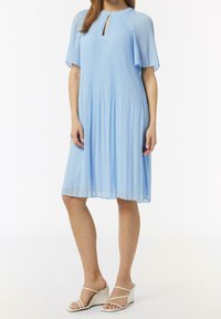 Himmelblaues plissiertes Kleid mit rundem Ausschnitt und Schlüsselaugen-Detail. Kurze Ärmel und eine fließende Silhouette. Getragen mit weißen Keilsandaletten.