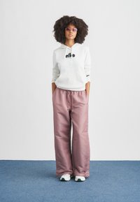 Hellgrauer Kapuzenpullover mit schwarzem Logo, kombiniert mit locker sitzenden pinken Hosen und weißen Sneakers mit grünen Akzenten. Überall glatte Texturen.