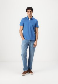 Blå polo skjorte med hvid krave, båret med lyseblå jeans og brune sneakers. Modellen står oprejst foran en almindelig hvid baggrund.