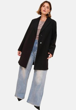 Zwarte oversized blazer met een reverskraag en twee knopen, gedragen over een taupe top, in combinatie met licht gewassen wide-leg jeans, en puntige zwarte schoenen.