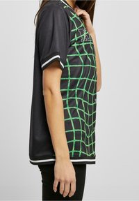Maglia nera con un motivo a griglia verde, maniche corte, colletto e orlo a strisce bianche. Presenta bottoni a pressione sul davanti.