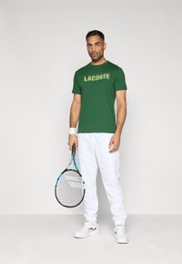 Camiseta de algodón verde con el logo "LACOSTE", pantalones deportivos blancos, raqueta de tenis y zapatillas; estilo deportivo casual.