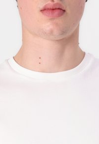 Fehér, kerek nyakú póló, puha anyagból készült, sima textúrával. A póló alatt két kicsi anyajegy látható a nyakon.