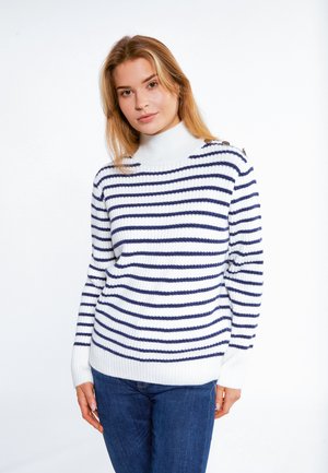 Ung kvinde med blondt hår iført en hvid rullekrave med marineblå vandrette striber og blå jeans, stående mod en hvid baggrund.