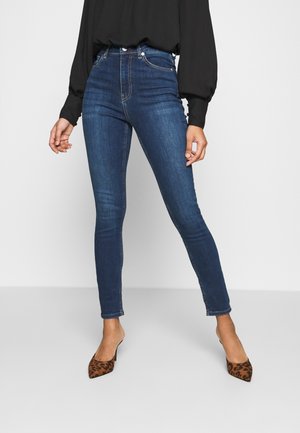 Kvinde iført mørkeblå højttaljede skinny jeans, sort langærmet bluse og leopardmønstrede spidse hæle står mod en ensfarvet baggrund.