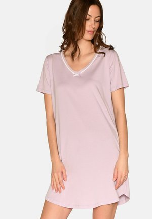 Frau trägt ein hellrosa Nachthemd mit kurzen Ärmeln, V-Ausschnitt und kleinem Schleifendetail, steht vor einem einfarbigen Hintergrund.