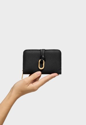 Mano sostiene una cartera de cuero negro con textura y cierre de cremallera y broche dorados sobre un fondo gris claro.