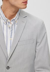 Blazer gris claro con un acabado texturizado, cierre de un solo botón y un bolsillo. Debajo, una camisa blanca con rayas verticales azules.