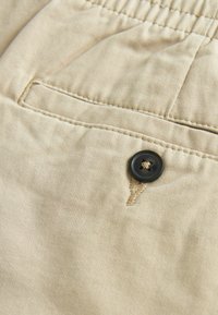 Gros plan sur un tissu beige avec une poche passepoilée et un bouton noir à quatre trous cousu près de la boutonnière cousue.