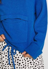 Klarblå stickad tröja med framficka, ribbade ärmslut och dragsko i fållen. Bärs över en vit klänning med svarta prickar.