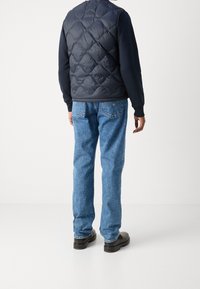 Marineblauer, gesteppter Puffer-Westen über einem schwarzen Langarmhemd, kombiniert mit hellblauen Jeans und schwarzen Schuhen, von hinten betrachtet.