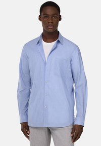 Camicia azzurra chiara a maniche lunghe con righe bianche verticali, chiusura a bottoni sul davanti, taschino sul petto e colletto aperto. Tessuto misto cotone.