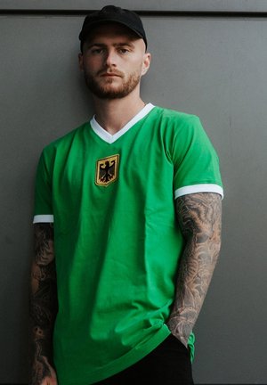 Grünes T-Shirt mit kurzen Ärmeln, weißen Akzenten und einem goldenen Emblem mit einem Adler. Weicher Stoff, lockere Passform und mit einem V-Ausschnitt-Design.