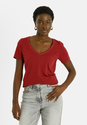 Femme aux cheveux courts et bouclés portant des boucles d'oreilles créoles en or, un T-shirt rouge à col en V et un jean gris clair, une main dans la poche.
