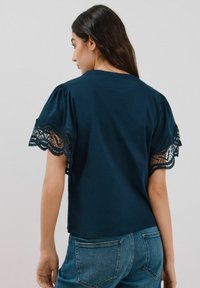 Dunkelblaues T-Shirt mit kurzen Puffärmeln, das eine detaillierte Spitzenborte aufweist. Der Stoff ist weich und glatt und zeigt eine lässige Passform.