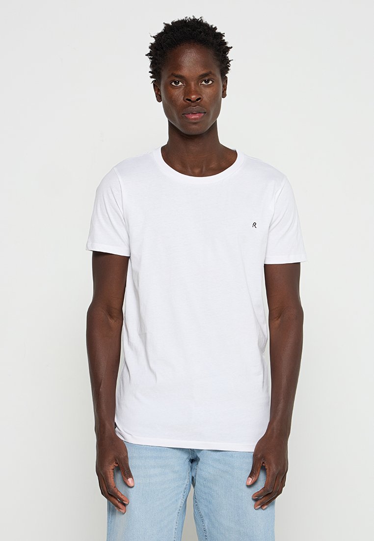 Biała bawełniana koszulka z okrągłym dekoltem i małym czarnym logo na piersi. Zestawiona z jasnoniebieskimi dżinsami.