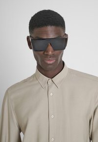 Gafas de sol negras de gran tamaño con lentes angulares y acentos metálicos. Llevadas con una camisa beige clara de botones hecha de una tela suave.