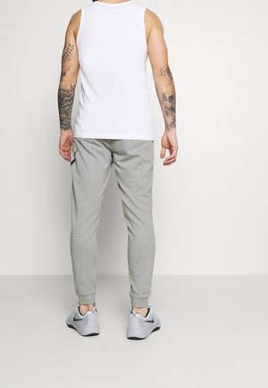 Weißes ärmelloses Tanktop, graue tapered Jogginghose und graue Sportschuhe mit schwarzen Akzenten. Haut mit sichtbaren Tattoos an den Armen.