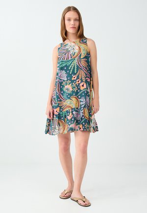 Femme debout portant une robe sans manches, longueur genou, avec des motifs floraux colorés et paisley, et des sandales plates beiges.