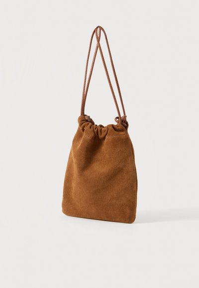 Petit sac bourse en daim marron avec cordon de serrage et fines bretelles suspendu sur un fond blanc uni.