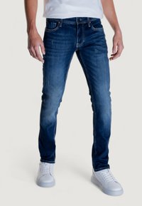 Jeans de mezclilla oscuros de corte ajustado con un desvanecimiento sutil, cinco bolsillos y costuras en contraste, combinados con zapatillas blancas y una camiseta blanca lisa.