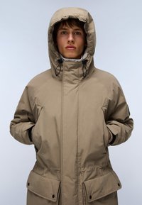 Beige isoleret parka med en stor hætte, lynlås foran og sidelommer. Har en glat tekstur og justerbare snore i kraven.