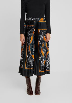 Kvinde iført en sort langærmet top, et plisseret midilangt skørt med orange, sort og hvide kædemønstre samt brune ankelstøvler.