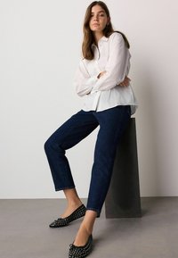 White button-up shirt with long sleeves, paired with dark blue denim jeans and black studded ballet flats, sitting on a black pedestal.