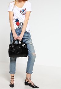 Bílý grafický triko s barevnými nášivkami, obnošené modré džíny, lesklá černá kabelka s kulatým detailem a černé šněrovací baleríny.