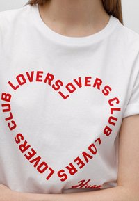 Hvid bomulds t-shirt med et rødt cirkulært tekstdesign, der står "LOVERS CLUB" og en mindre tekst "Huac" nederst. Korte ærmer.