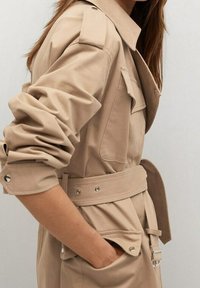 Femme aux longs cheveux portant un trench-coat beige ceinturé avec des manches retroussées et des poches à rabat à l'avant, la main dans la poche, sur un fond uni.