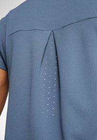 Modrá košile s krátkým rukávem z texturované látky. Má záševky na zádech, které odhalují síťovanou část s tečkovanými detaily pro ventilaci.