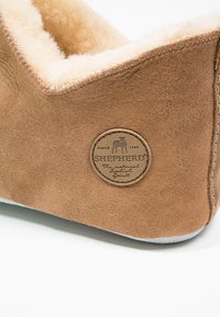 Chausson en daim marron avec doublure en fourrure crème, empiècement en cuir rond affichant le nom de la marque et des coutures le long du côté.